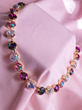 A gold-tone necklace with round iridescent crystals in pink, purple, and multicolor tones, displayed on a draped light pink fabric background.