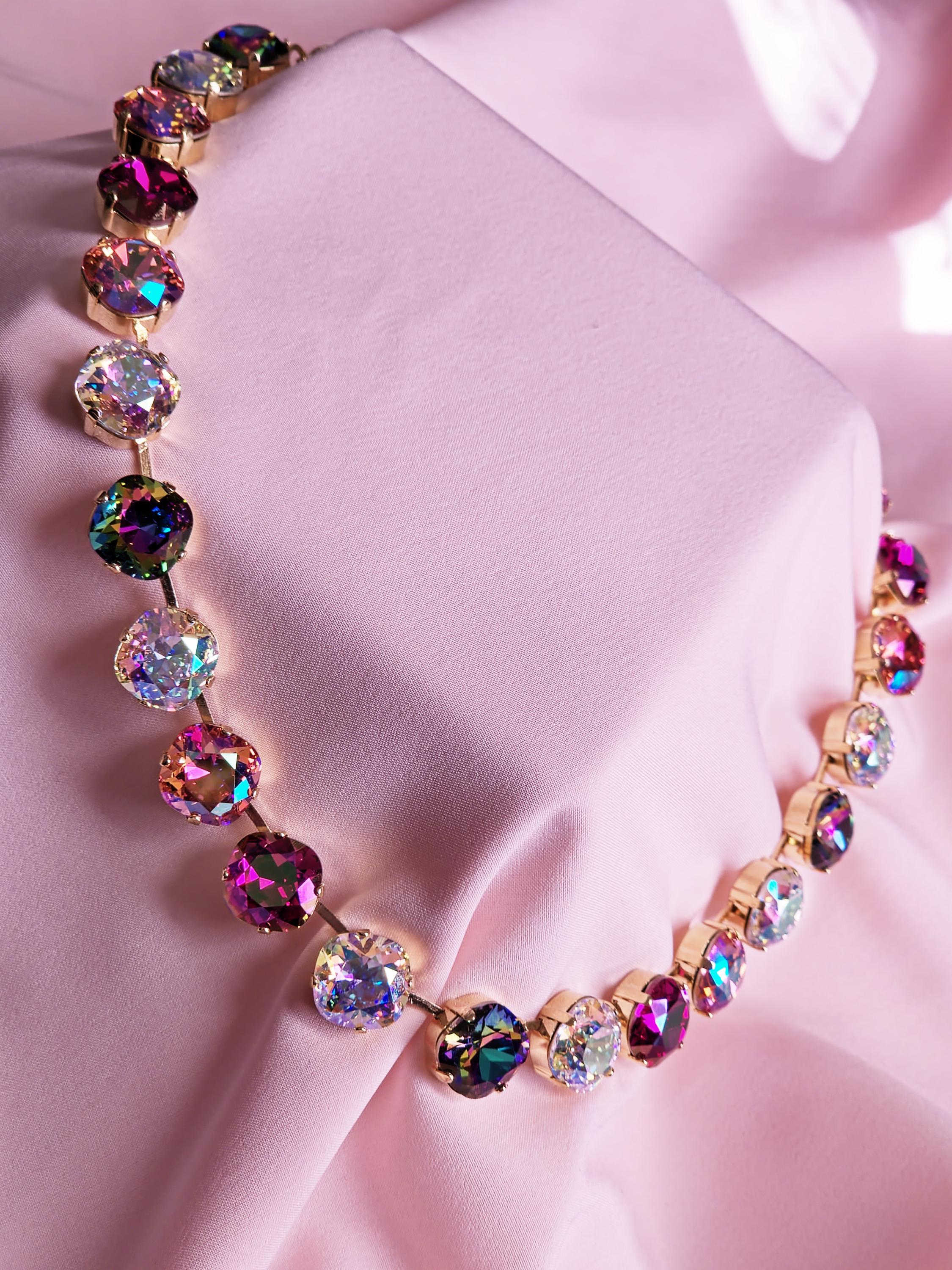 A gold-tone necklace with round iridescent crystals in pink, purple, and multicolor tones, displayed on a draped light pink fabric background.